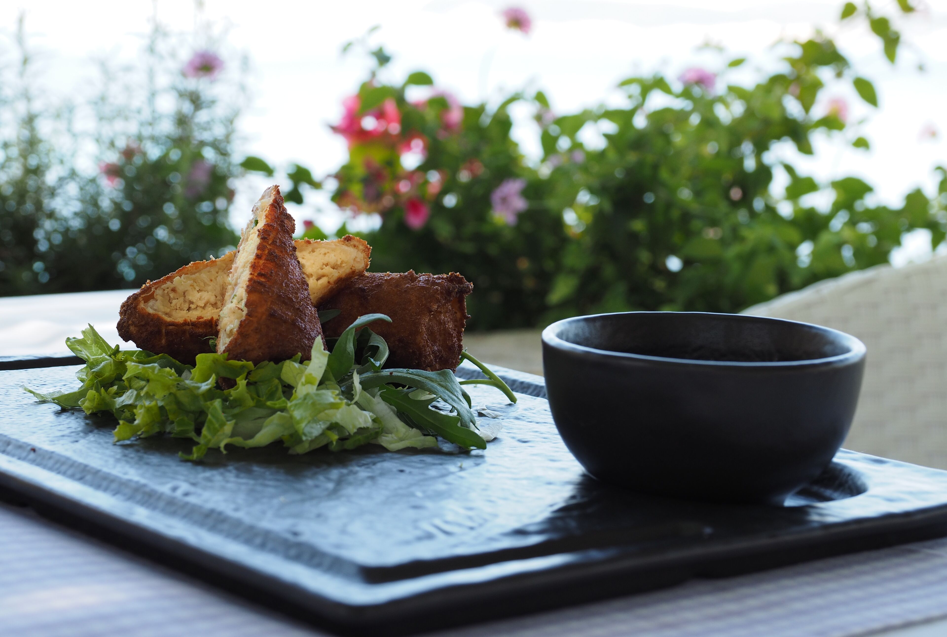 Un repas composé de pain et de légumes verts, servi sur une planche d’ardoise, accompagné d’un bol noir, le tout disposé en extérieur, avec de la verdure et des fleurs en arrière-plan.