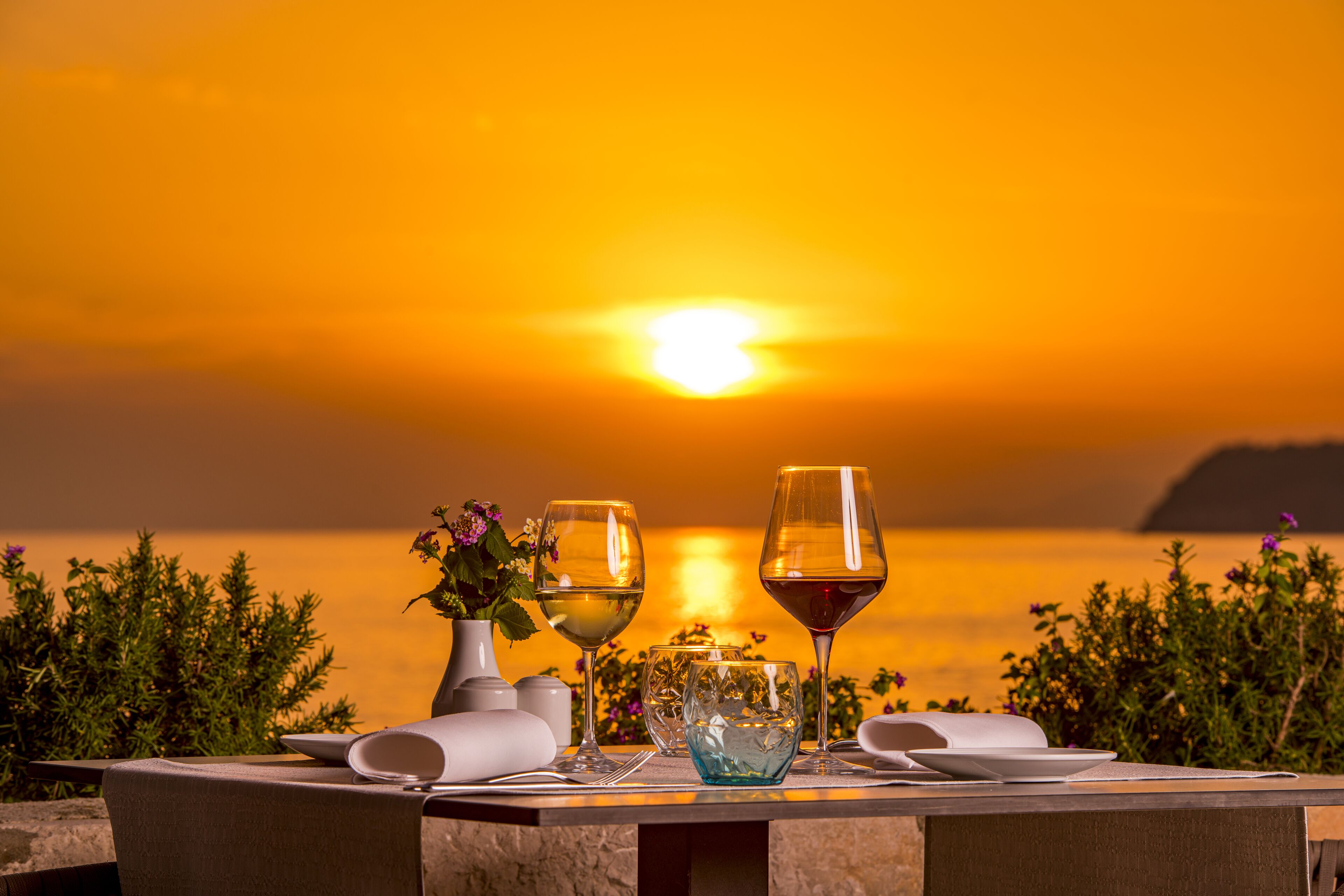 Mesa para dos con copas de vino y un jarrón de flores, con vista a un atardecer sobre el agua.
