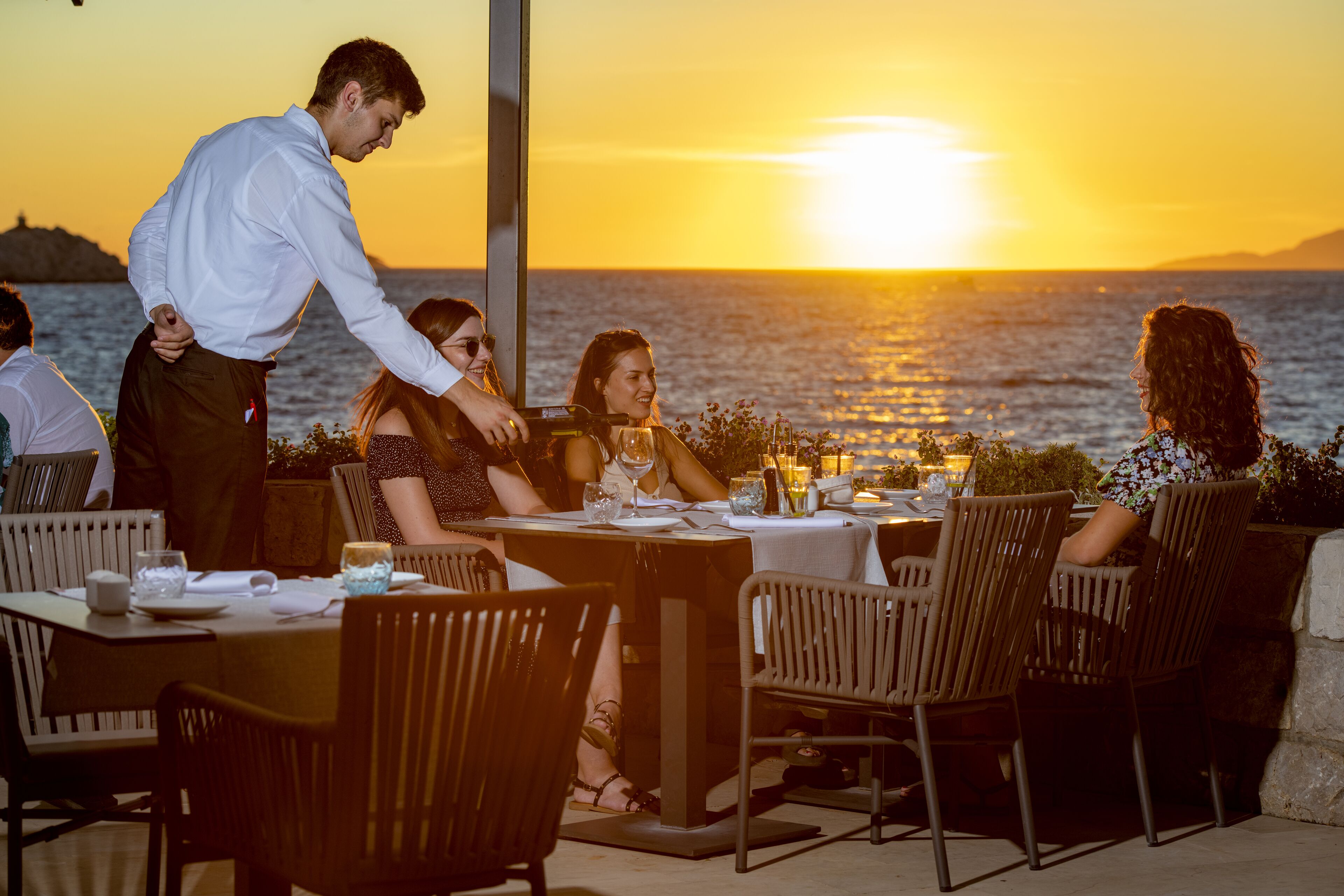 A restaurant with outdoor seating overlooking the sea, tables set with glasses and cutlery, and a waiter attending to guests.
