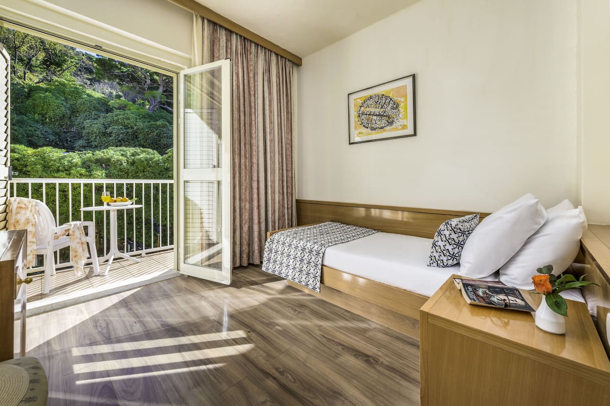 A hotel room with a bed, a small table with a vase, a chair, and a balcony with a view of greenery.
