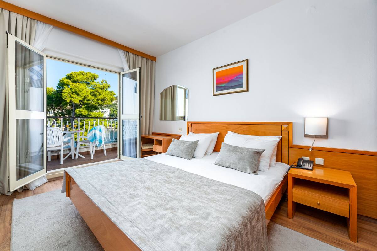 A hotel room with a large bed, a wooden headboard, a bedside table with a lamp, and a view of the outdoors through a sliding glass door.