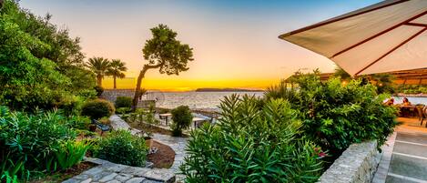 A garden with stone pathways, lush greenery, and outdoor seating overlooking a body of water at sunset.