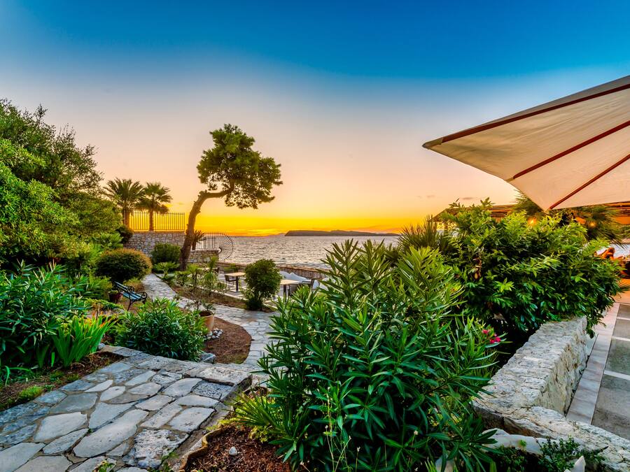 A garden with stone pathways, lush greenery, and outdoor seating overlooking a body of water at sunset.