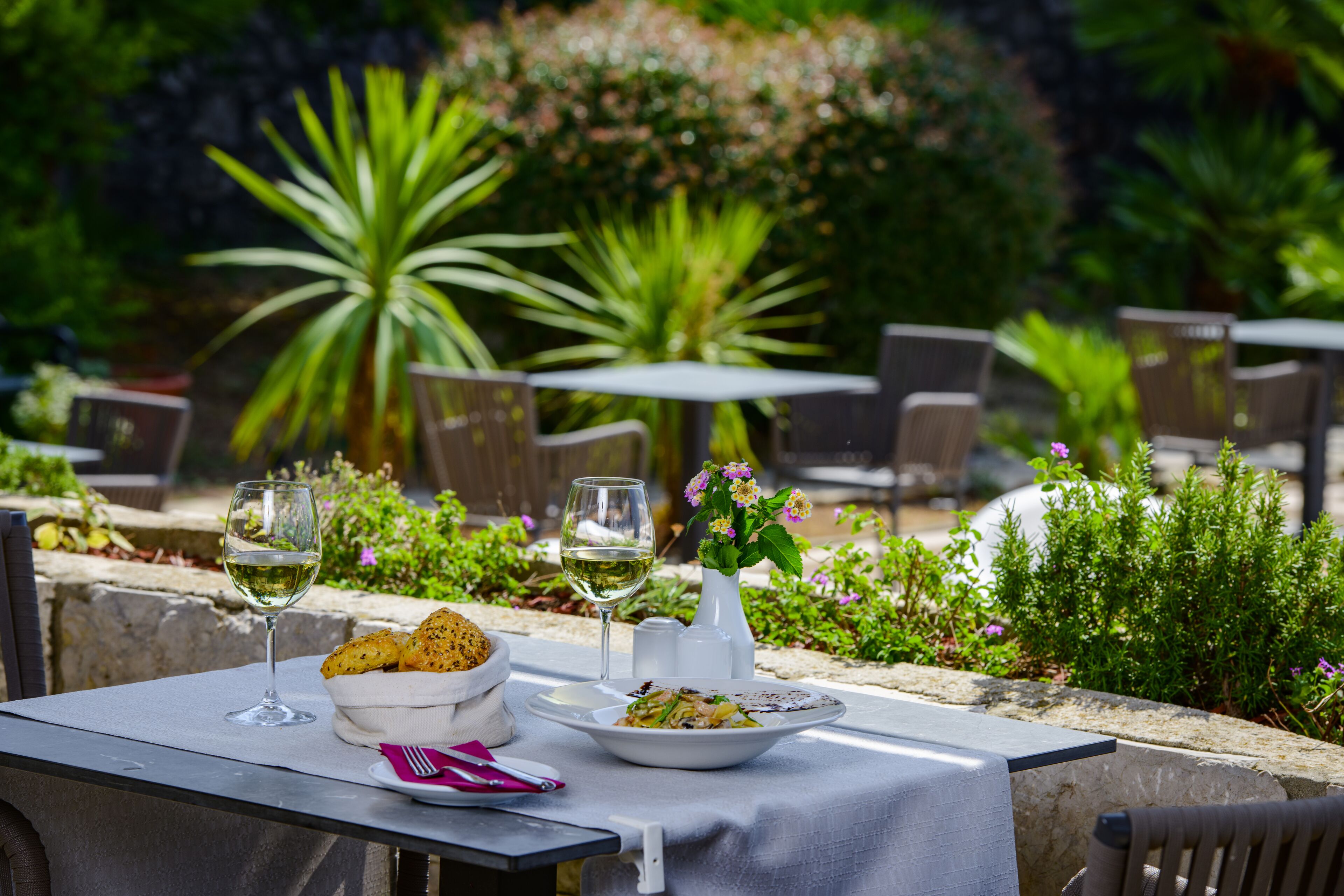 An outdoor dining area with a table set for two, featuring a bowl of bread, a plate of food, and two glasses of white wine.