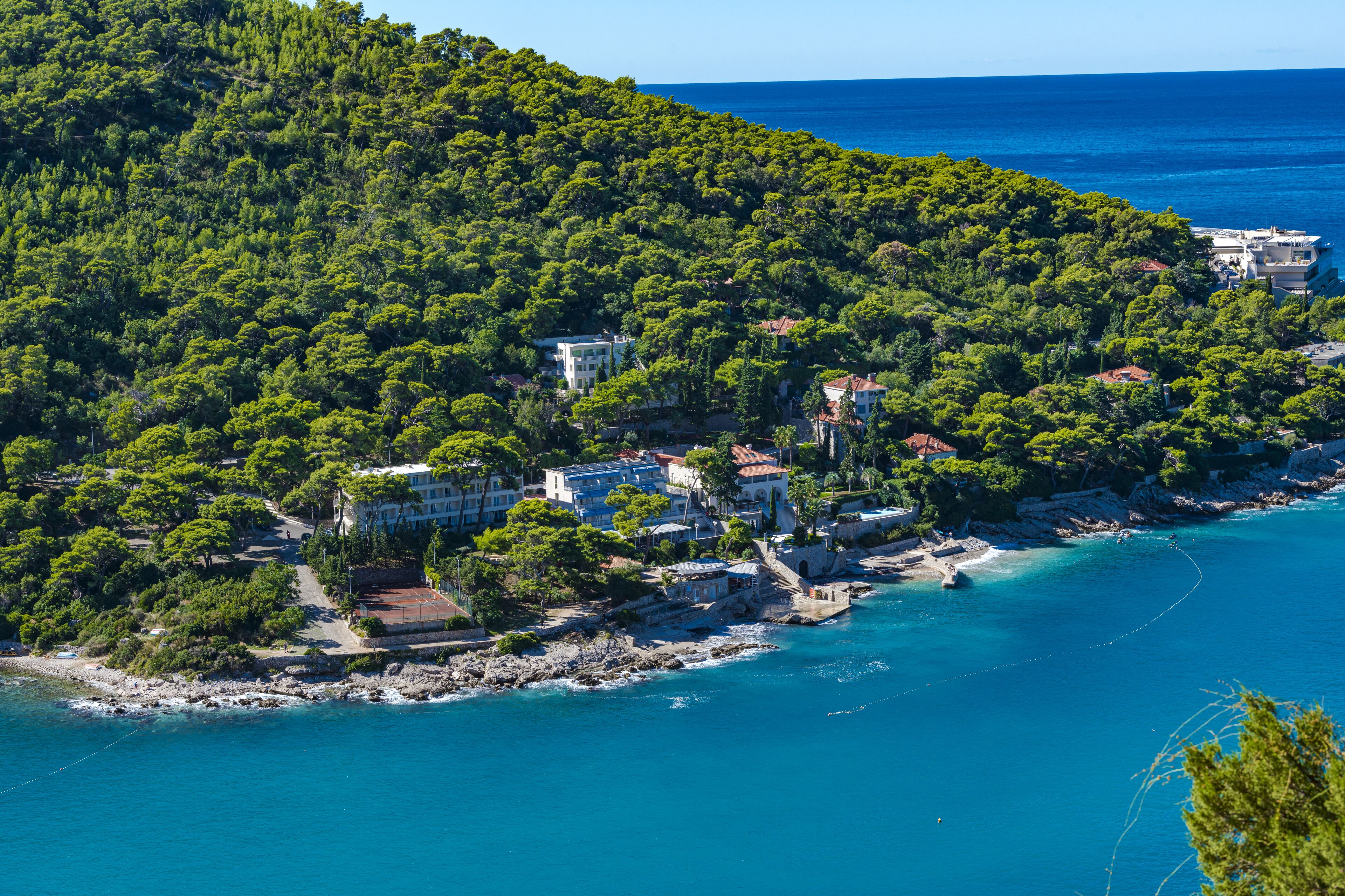 An aerial view of a coastal area with buildings surrounded by dense green forest, a clear blue sea, and rocky coastline.