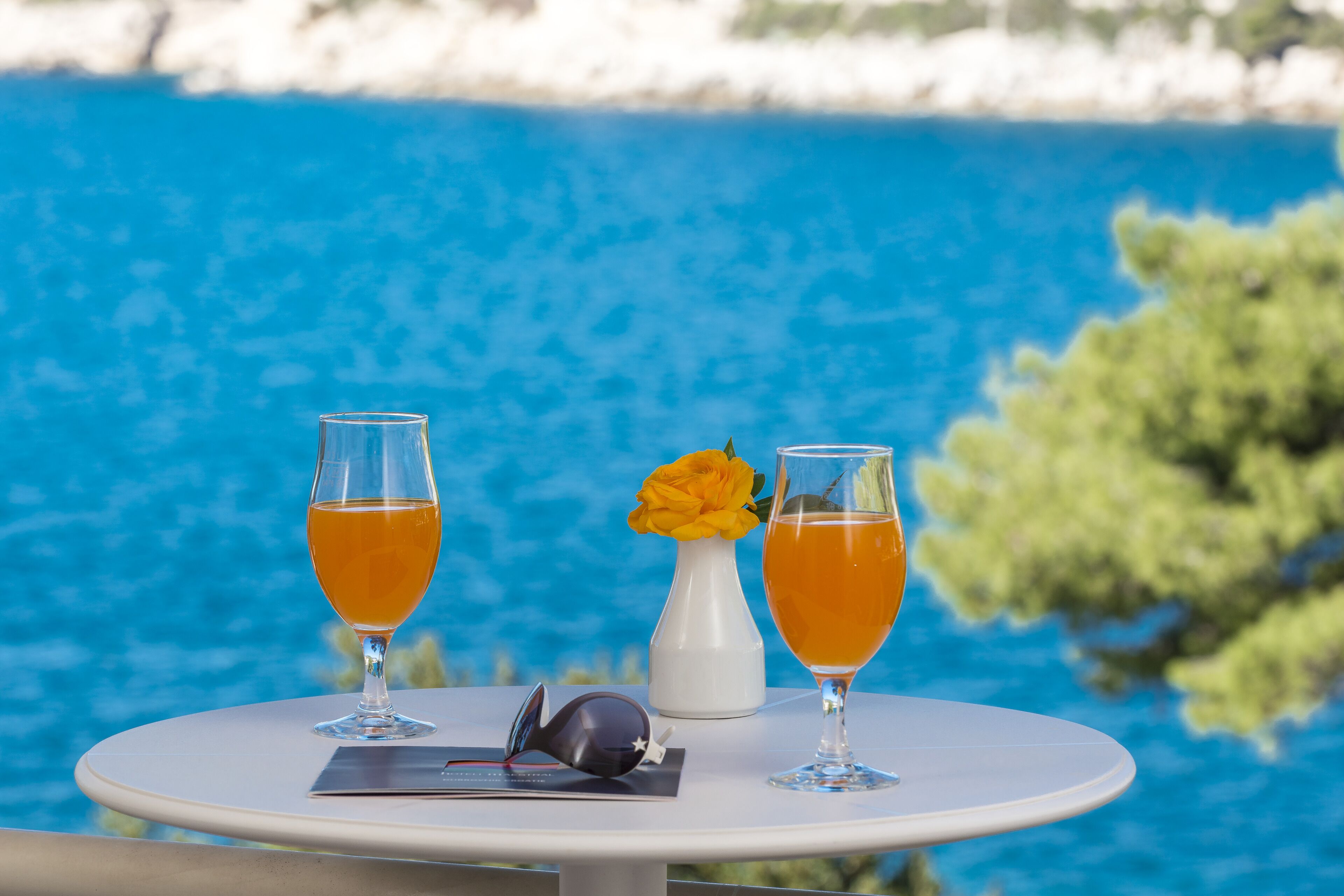 A table with two glasses of orange liquid, a white vase with a yellow flower, and a pair of sunglasses on a white table against a backdrop of a blue body of water and greenery.