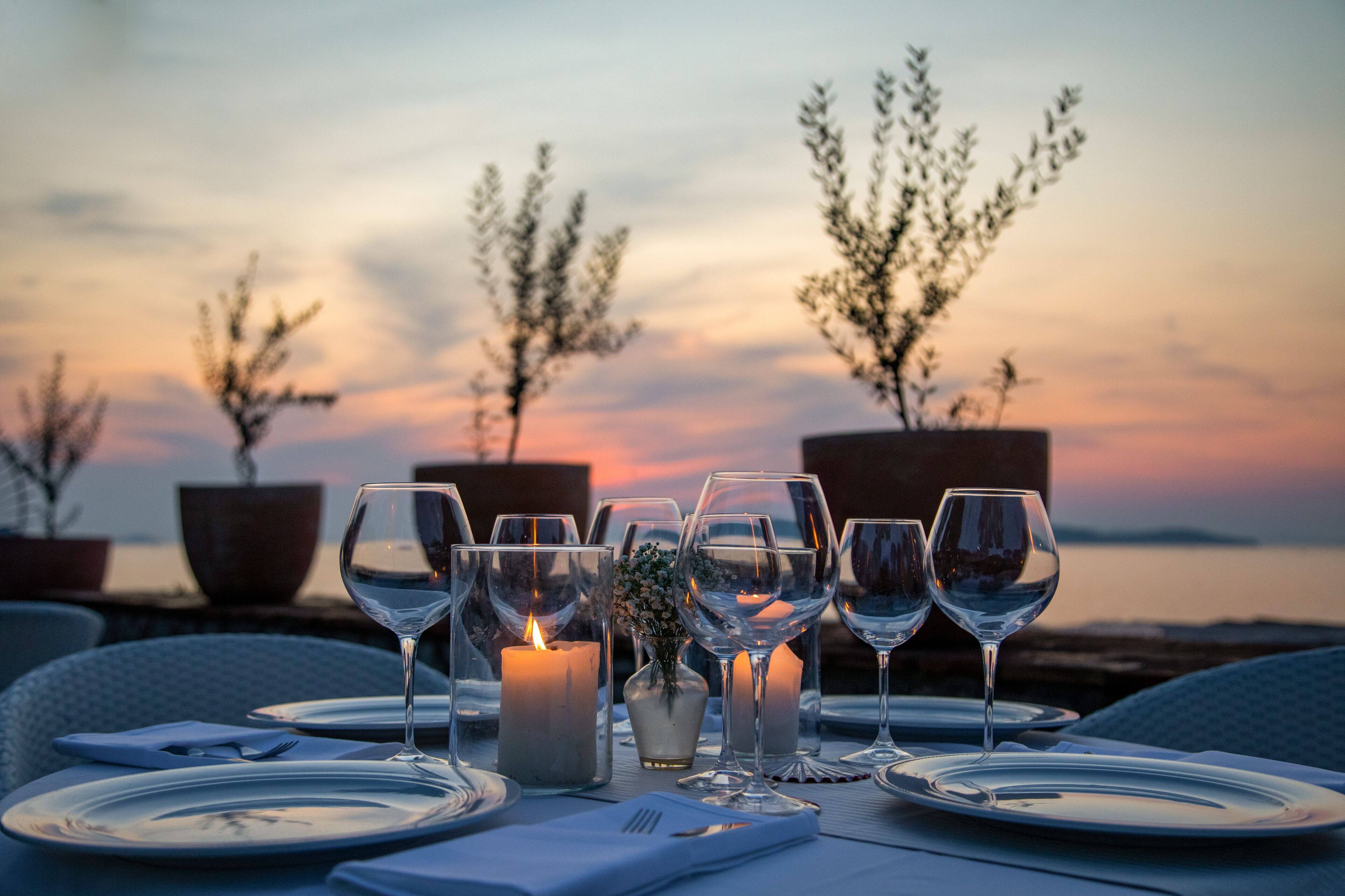 A table set for a meal with wine glasses, plates, and a candle, overlooking a sunset and trees.