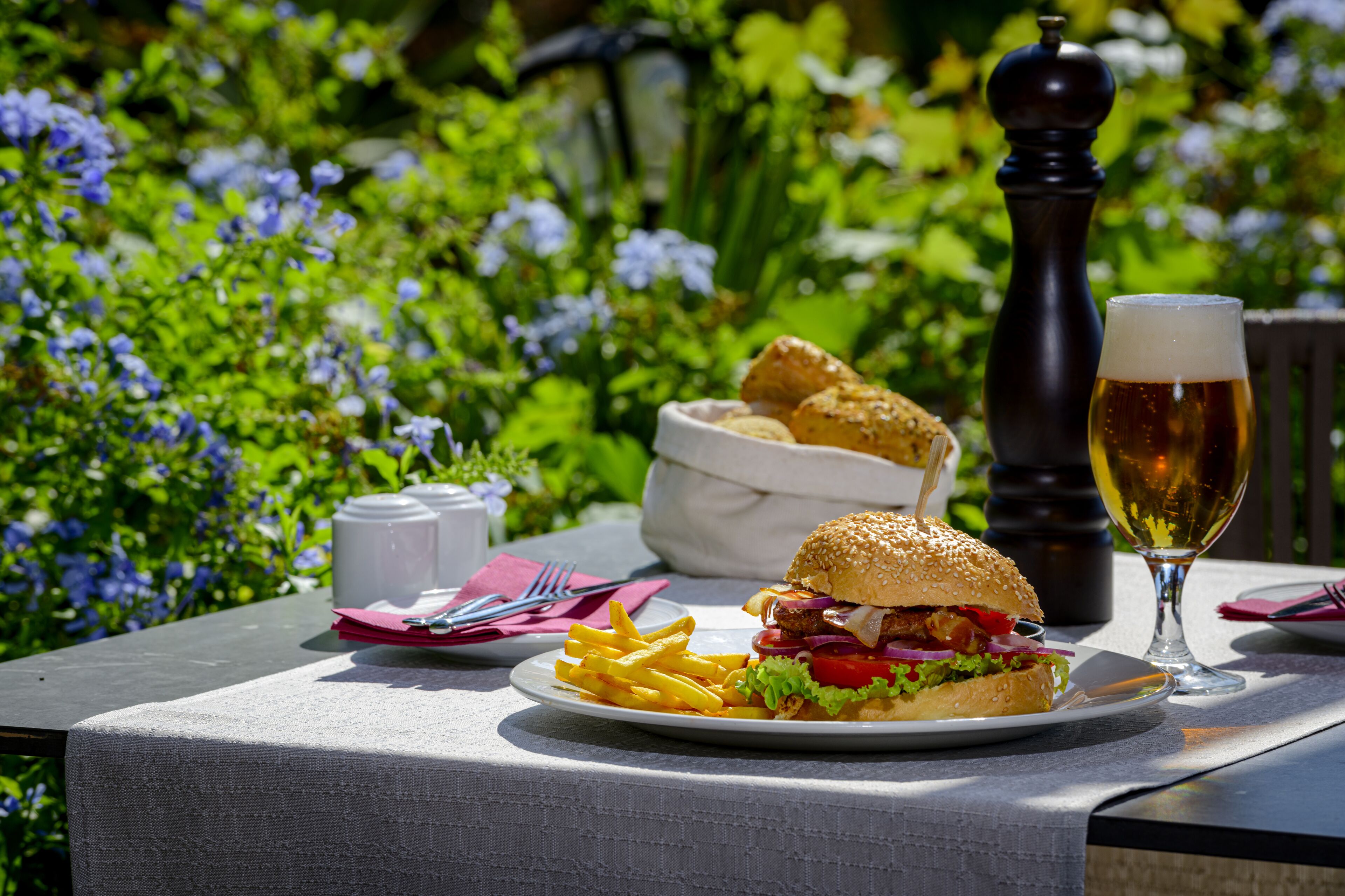 A meal with a burger and fries on a table outdoors, with a glass of beer and a pepper shaker in the background.