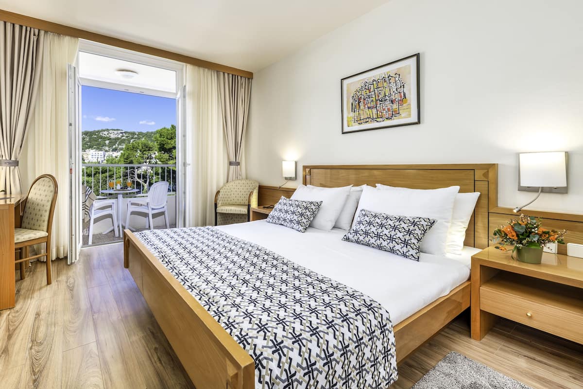 A hotel room with a large bed, a desk with a lamp, a chair, a small table with fruit, and a balcony with chairs and a table.