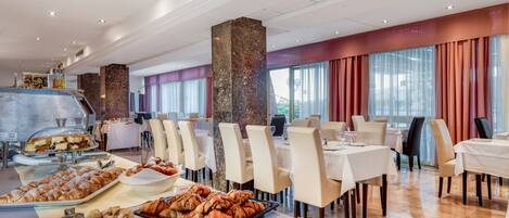 Area ristorazione in hotel con tavoli, sedie e un buffet con pasticcini e pane.