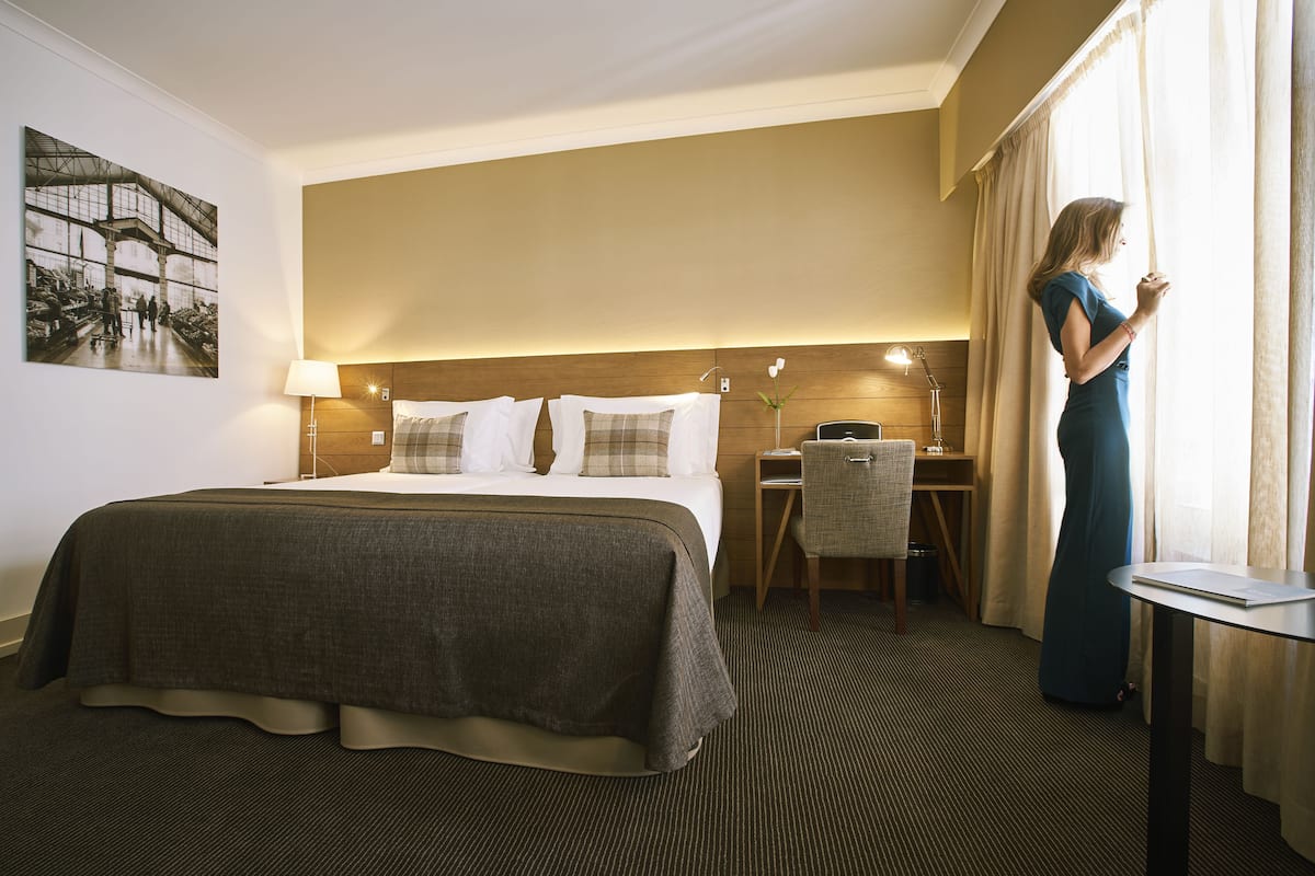 Quarto de hotel com uma cama grande, uma escrivaninha, uma cadeira e uma janela com cortinas.