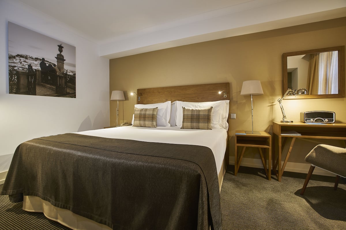 Quarto de hotel com uma cama grande, duas luminárias de cabeceira, uma escrivaninha de madeira com cadeira, um banquinho pequeno e um quadro na parede.