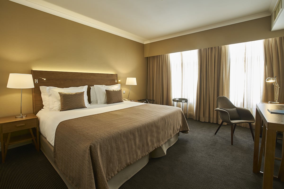 Quarto de hotel com uma cama grande, duas mesas de cabeceira com luminárias, uma escrivaninha com cadeira e uma janela ampla com cortinas.