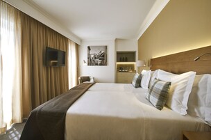 A hotel room with a large bed, a flat-screen TV mounted on the wall, a desk with a lamp, and a small seating area.