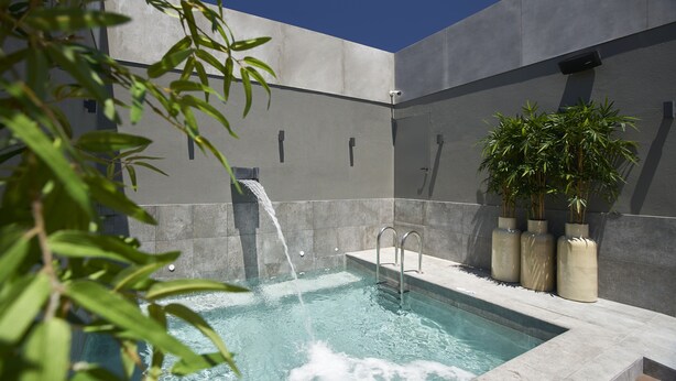 A modern outdoor pool with a waterfall feature, surrounded by stone walls and potted plants.