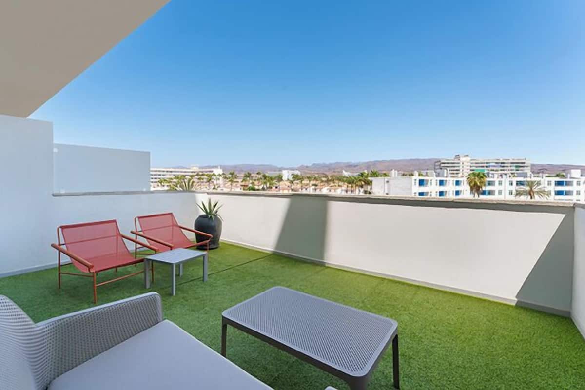 A rooftop terrace with artificial grass, two red lounge chairs, a small table, and a potted plant.