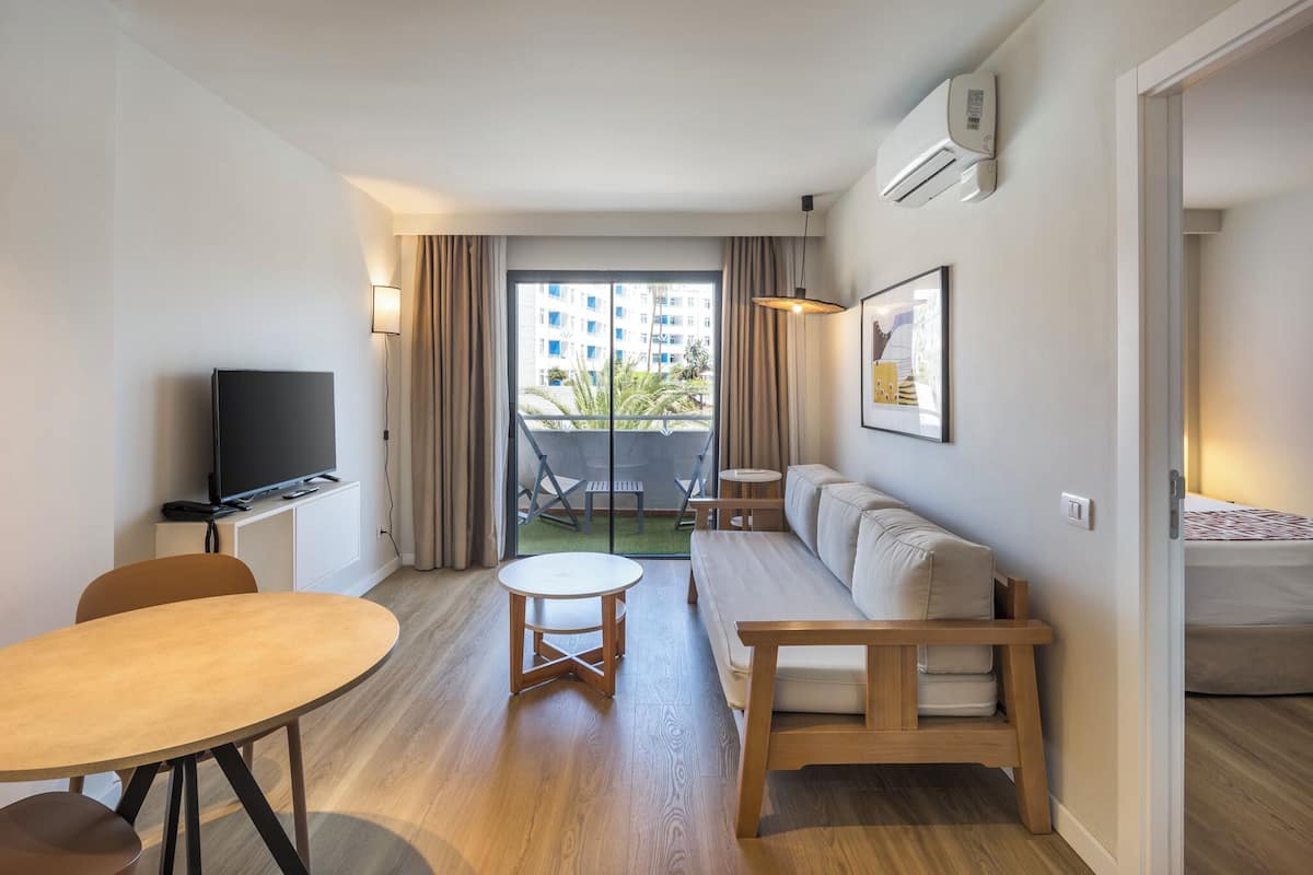 A modern hotel room with a sofa, a small wooden table, a TV, and a balcony with a view of a building and greenery.