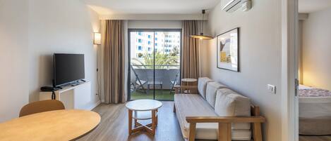 A modern hotel room with a flat-screen TV, a sofa, a small table, and a balcony with a view of a building and greenery.
