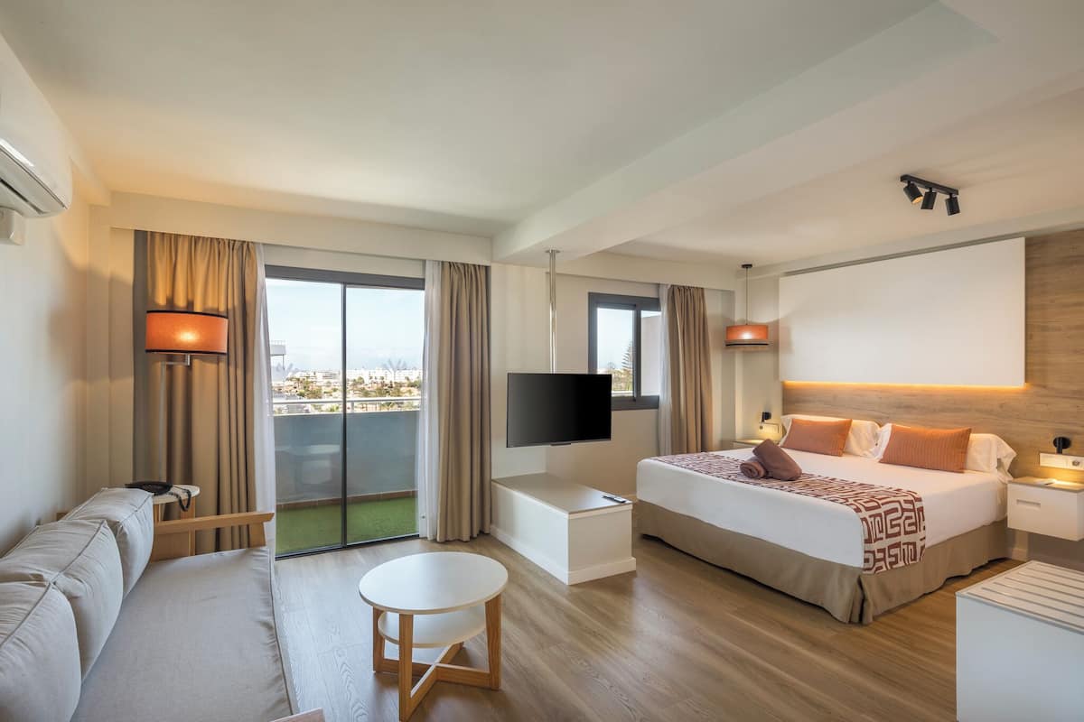 A modern hotel room with a bed, sofa, TV, and a balcony with curtains.