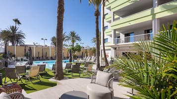 Zona junto a la piscina con sillones de descanso, palmeras y un edificio con balcones.