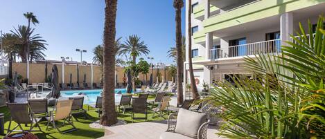Zona junto a la piscina con sillones de descanso, palmeras y un edificio con balcones.