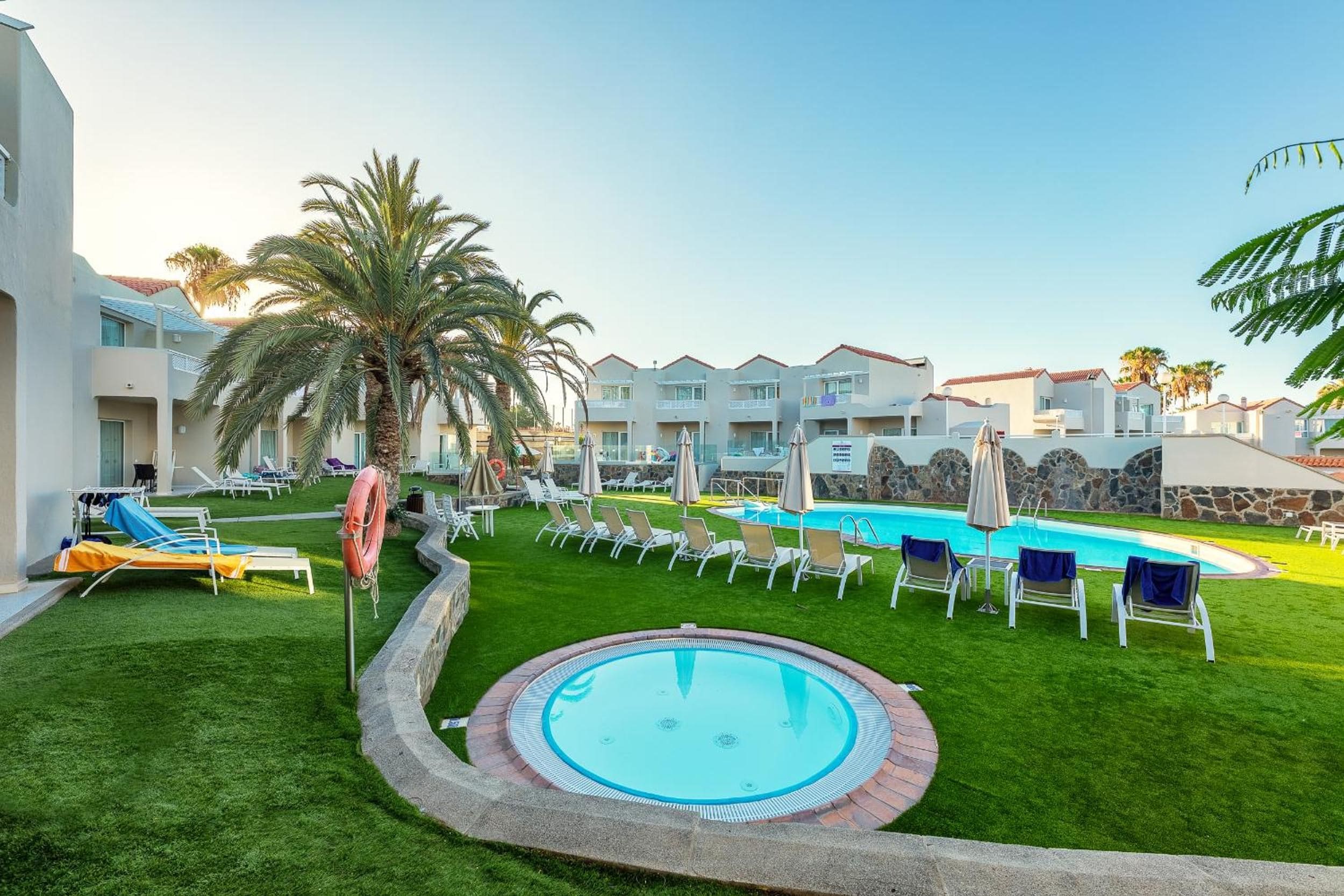 Un área de piscina con sillones de descanso y sombrillas, rodeada de césped cuidado y palmeras.