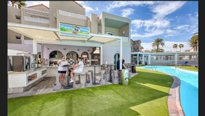 A poolside area with a bar and seating, a pool, and a building with a sign above the bar. - Apartamentos LIVVO Koala Garden (San Bartolomé de Tirajana)