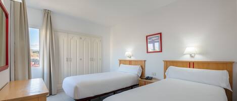 A hotel room with two single beds, a wooden nightstand, a desk, and a window with curtains.