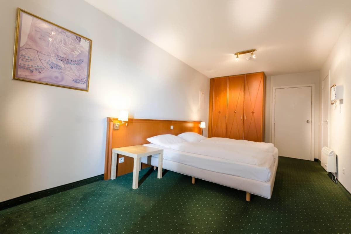 A hotel room with a single bed, a wooden wardrobe, a small table, and a wall-mounted light.