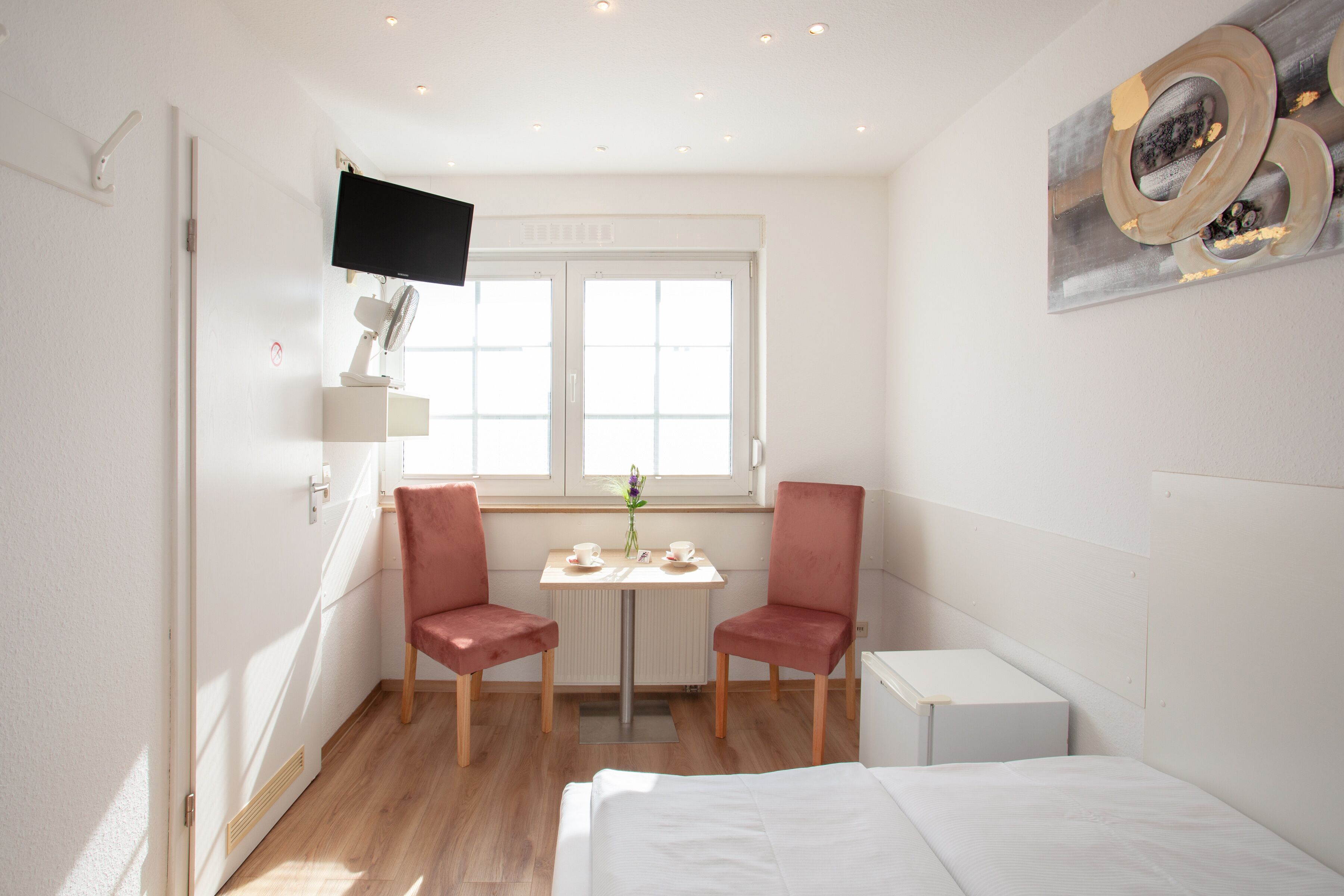 A hotel room with a bed, a desk with two chairs, a TV mounted on the wall, and a window with blinds.