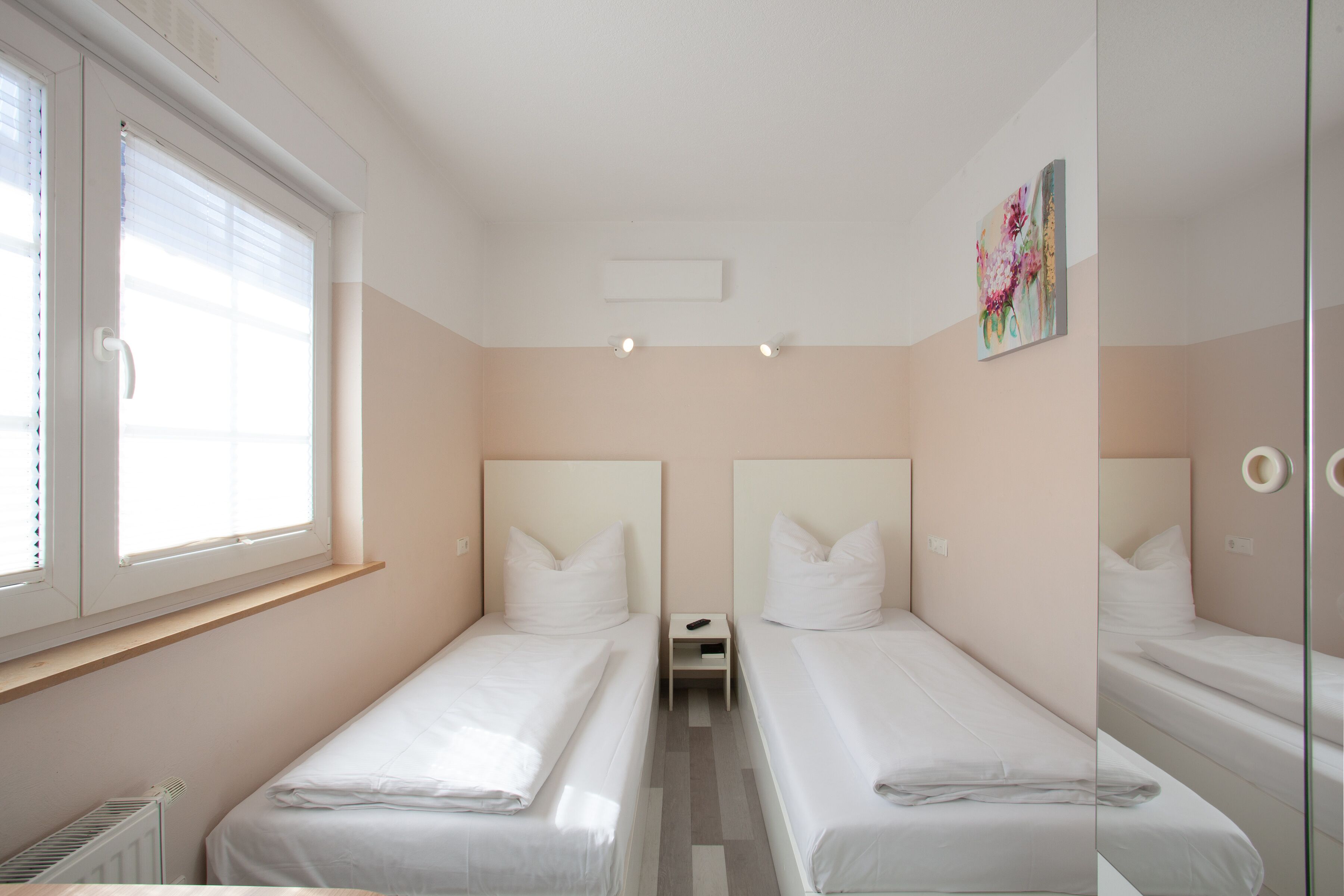 A bedroom with two single beds, a small bedside table, a radiator, and a window with blinds.