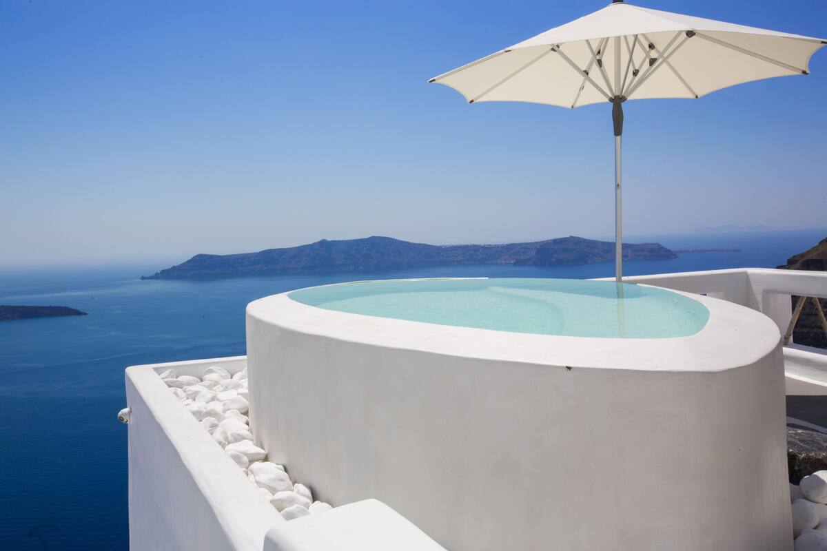 A circular infinity pool with a white umbrella, surrounded by white stone walls, overlooking a clear blue sea and distant islands.