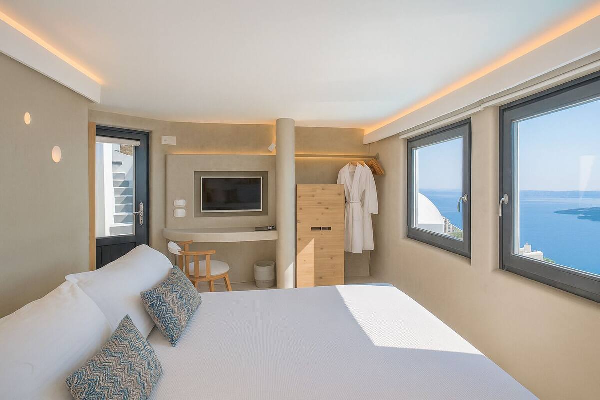 A modern hotel room with a large bed, a desk, a TV, and a view of the sea.