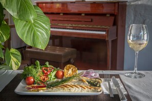 A dining table with a plate of food, a glass of white wine, and a piano in the background. - Flora Hotel Bodrum  (Bodrum)