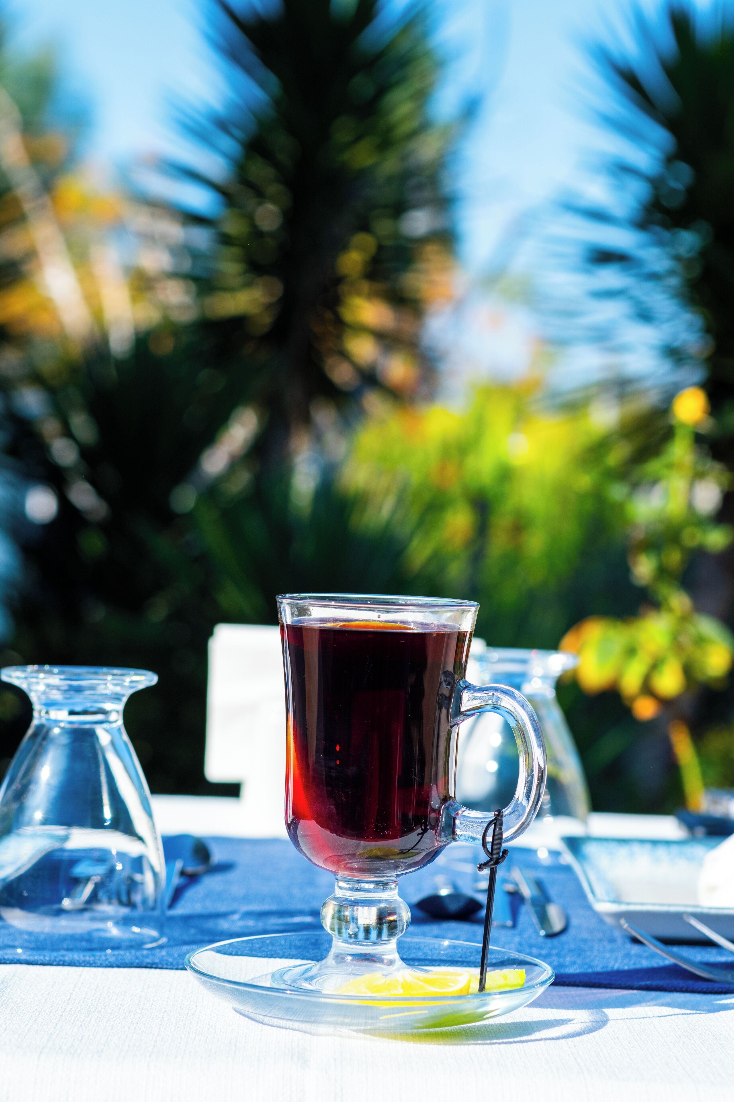 Una taza de té transparente sobre una mesa con mantel azul, rodeada de botellas de vidrio transparente y un paño blanco.