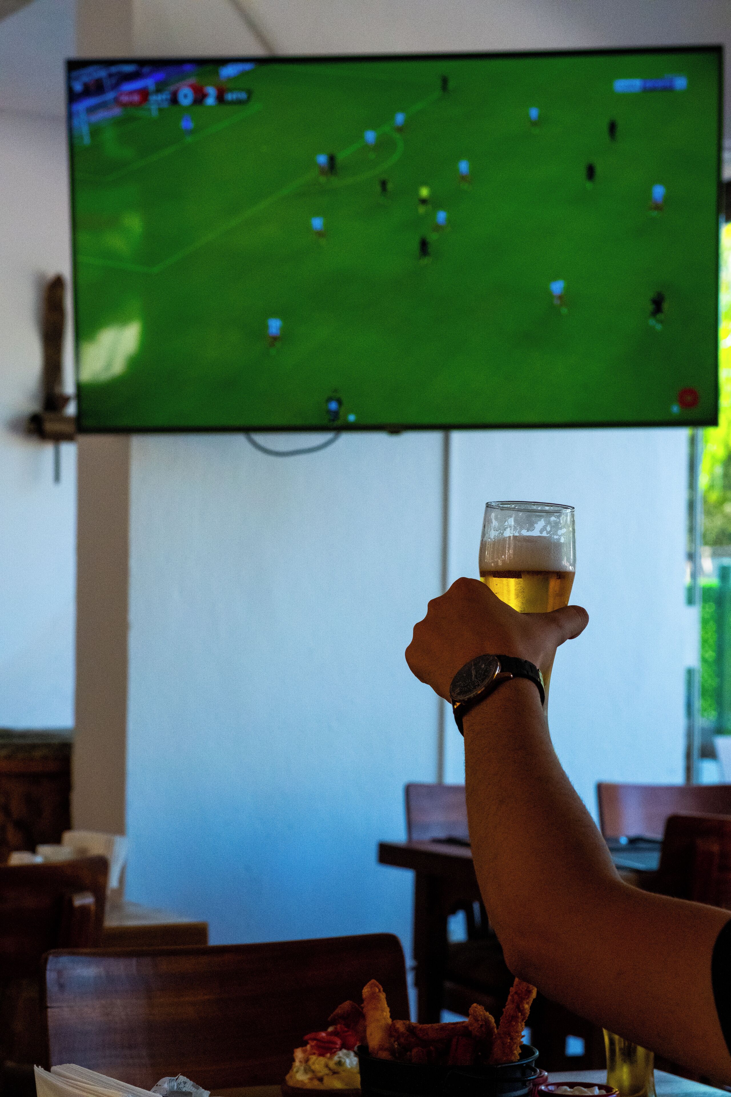 Una mano sosteniendo una jarra de cerveza frente a un televisor que muestra un partido de fútbol.