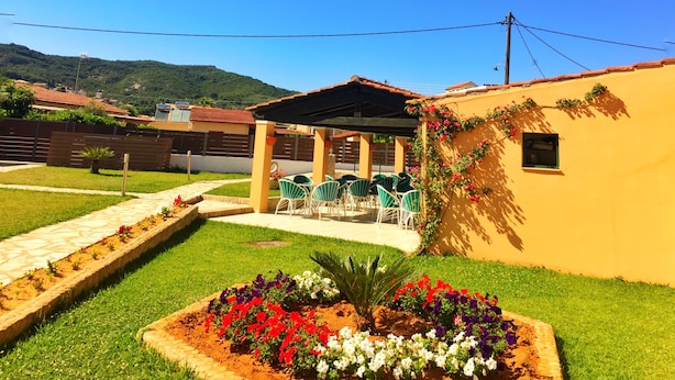 A hotel with a covered outdoor seating area, a garden with colorful flowers, and a view of a hill in the background. - Hotel Sea Bird (Corfu)