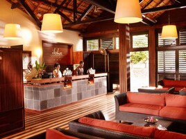 Un área de recepción con un mostrador de madera, un espacio de espera con mobiliario de mimbre y ventanas amplias con contraventanas de madera.