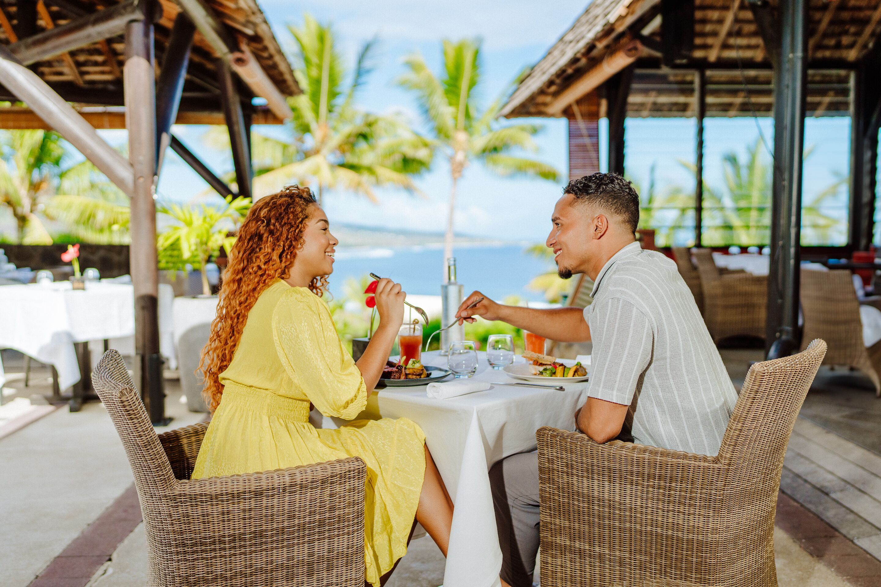 Un couple est assis à une table en plein air, profitant de la vue sur l’océan et les palmiers.