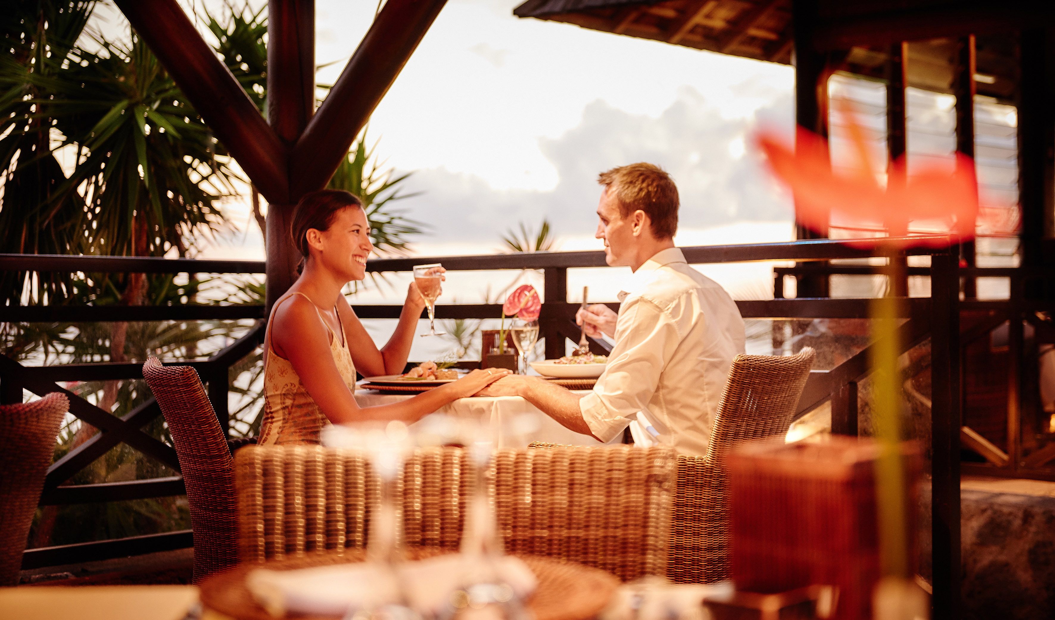 Un couple est assis à une table à l’extérieur, sur une terrasse meublée de mobilier en osier, et profite d’une vue panoramique.