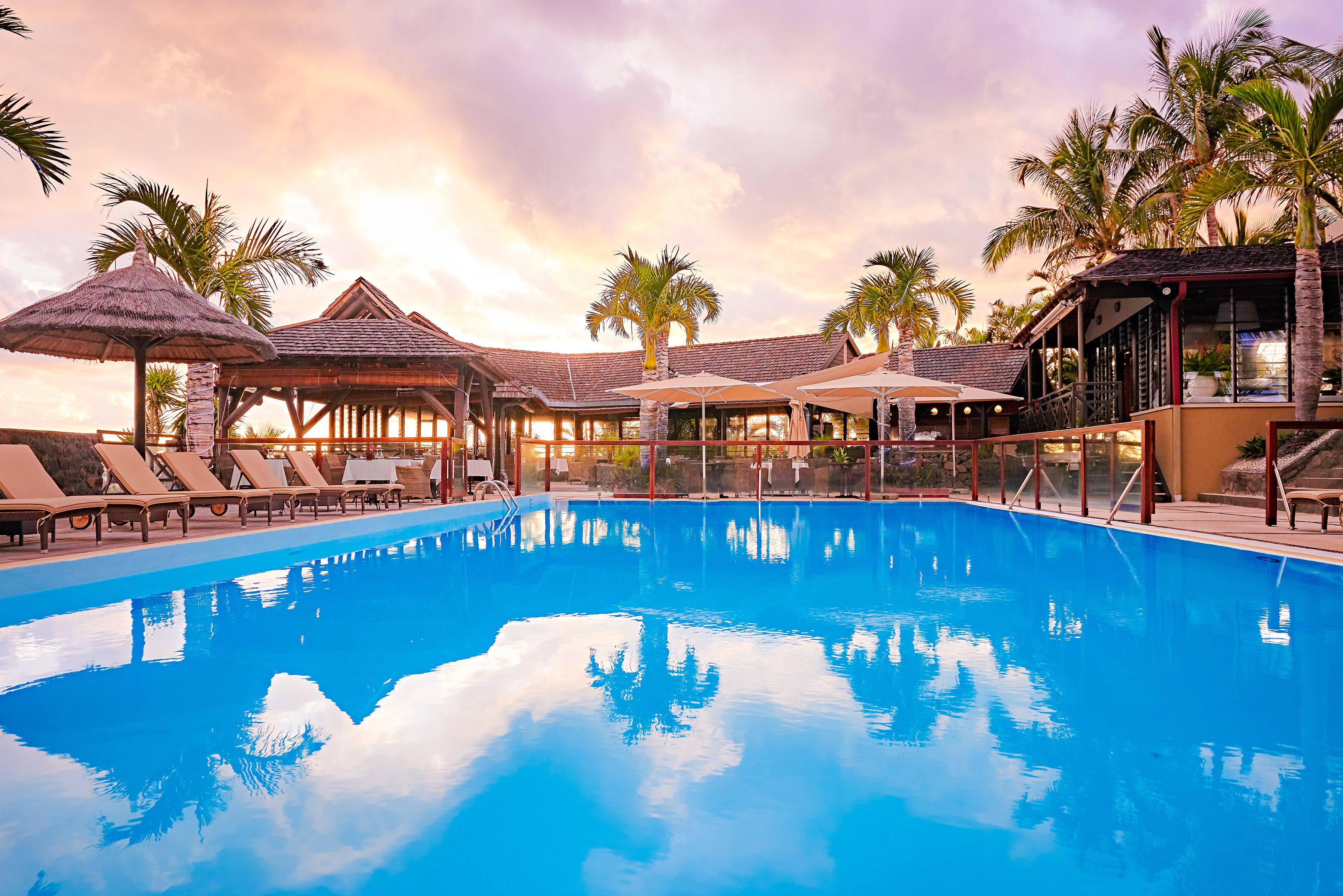 A swimming pool with lounge chairs and umbrellas, surrounded by palm trees and covered structures.