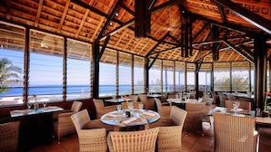 Un restaurante con techo de madera y amplios ventanales con vista al océano. Las mesas están dispuestas con sillas y cubertería.