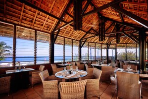 Un restaurante con techo de madera y amplios ventanales con vista al océano. Las mesas están dispuestas con sillas y cubertería.