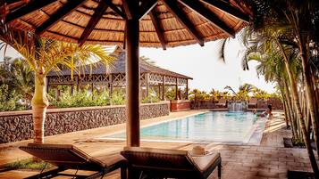 A pool area with lounge chairs, a thatched roof structure, and a stone wall.