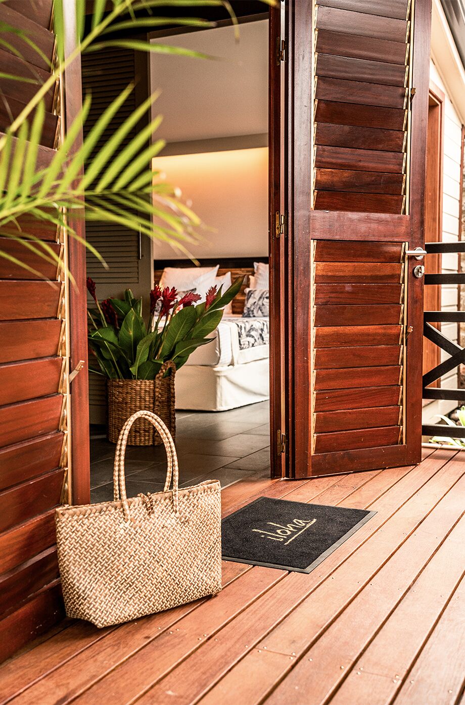 A wooden door with a doormat, a woven bag, and a potted plant on a wooden deck.