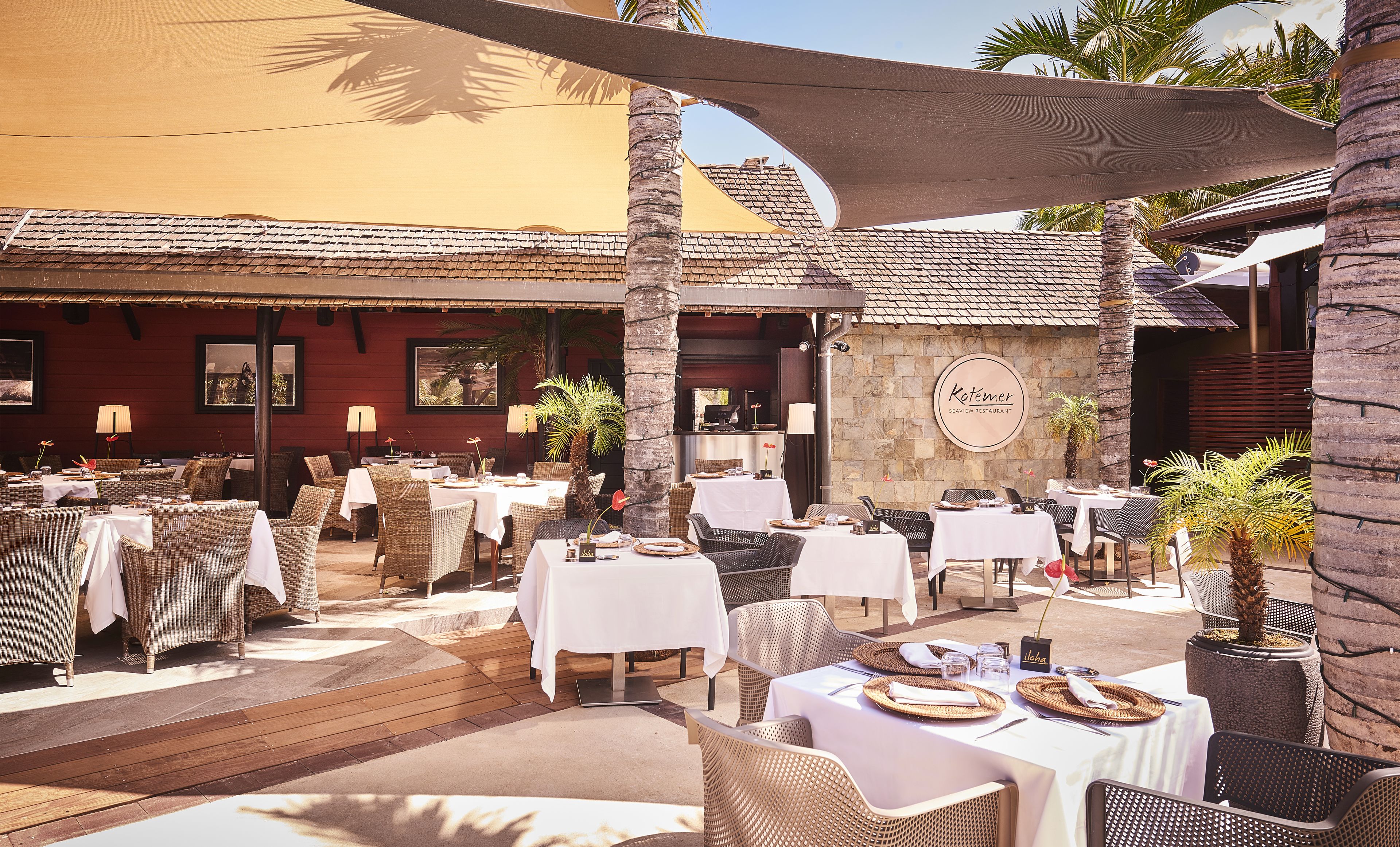 An outdoor restaurant with wicker furniture, white tablecloths, and a stone wall with a sign.