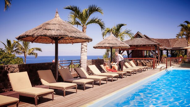 Zona de piscina con sillas de descanso y sombrillas de paja, con vista al mar azul y transparente.