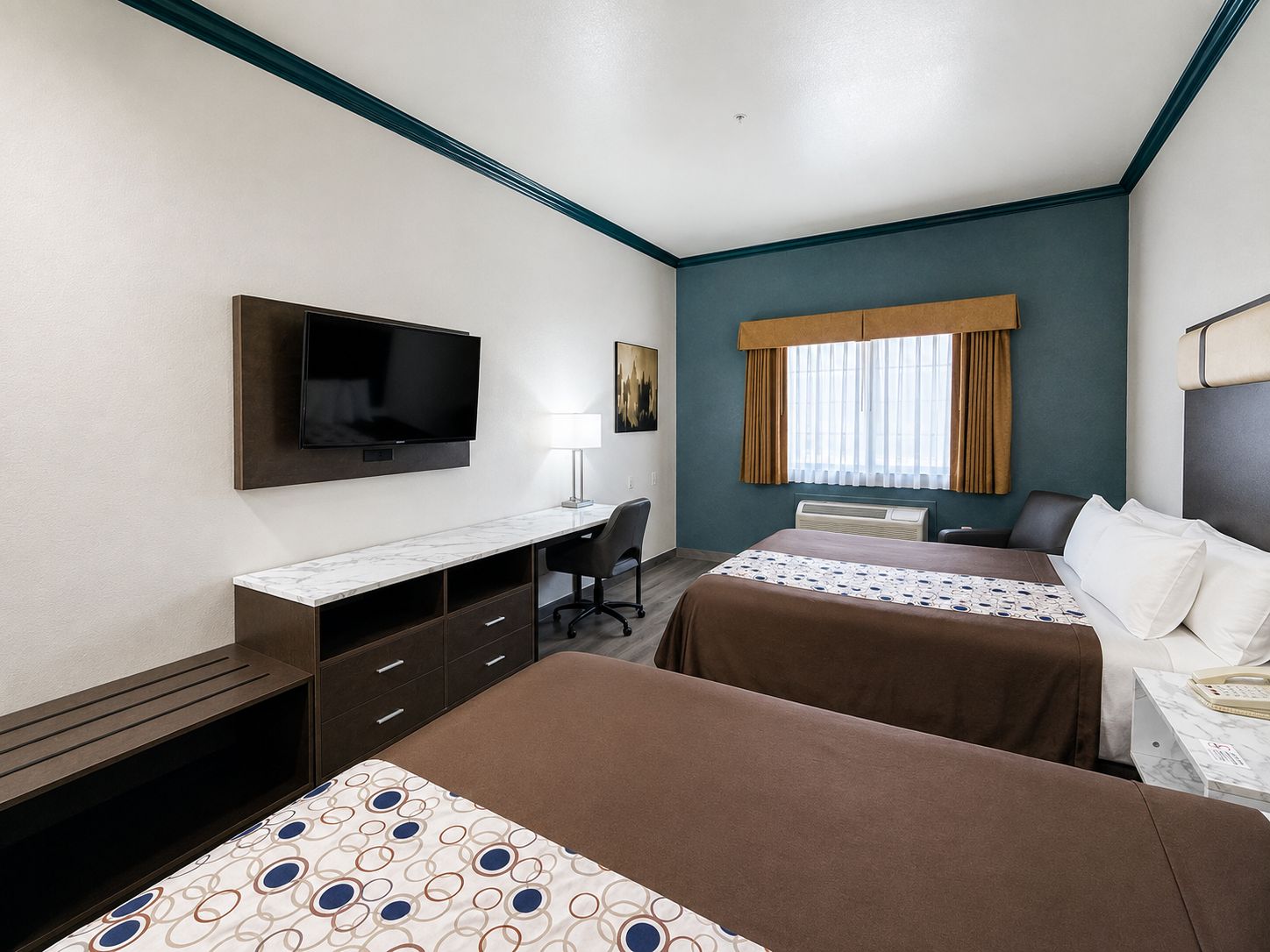 A hotel room with a flat-screen TV, a desk, two beds, a window with curtains, and a wall-mounted air conditioning unit.