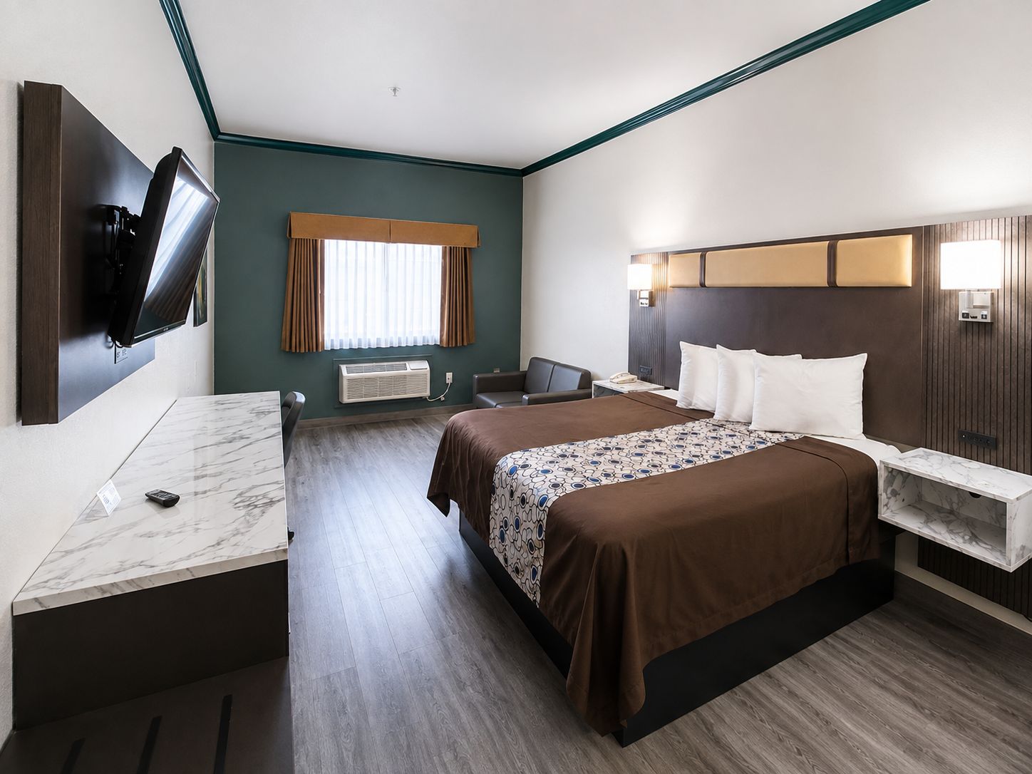 Ett hotellrum med en säng, sängbord, ett skrivbord, en platt-TV som är monterad på väggen, ett fönster med gardiner och en väggmonterad luftkonditioneringsanläggning.