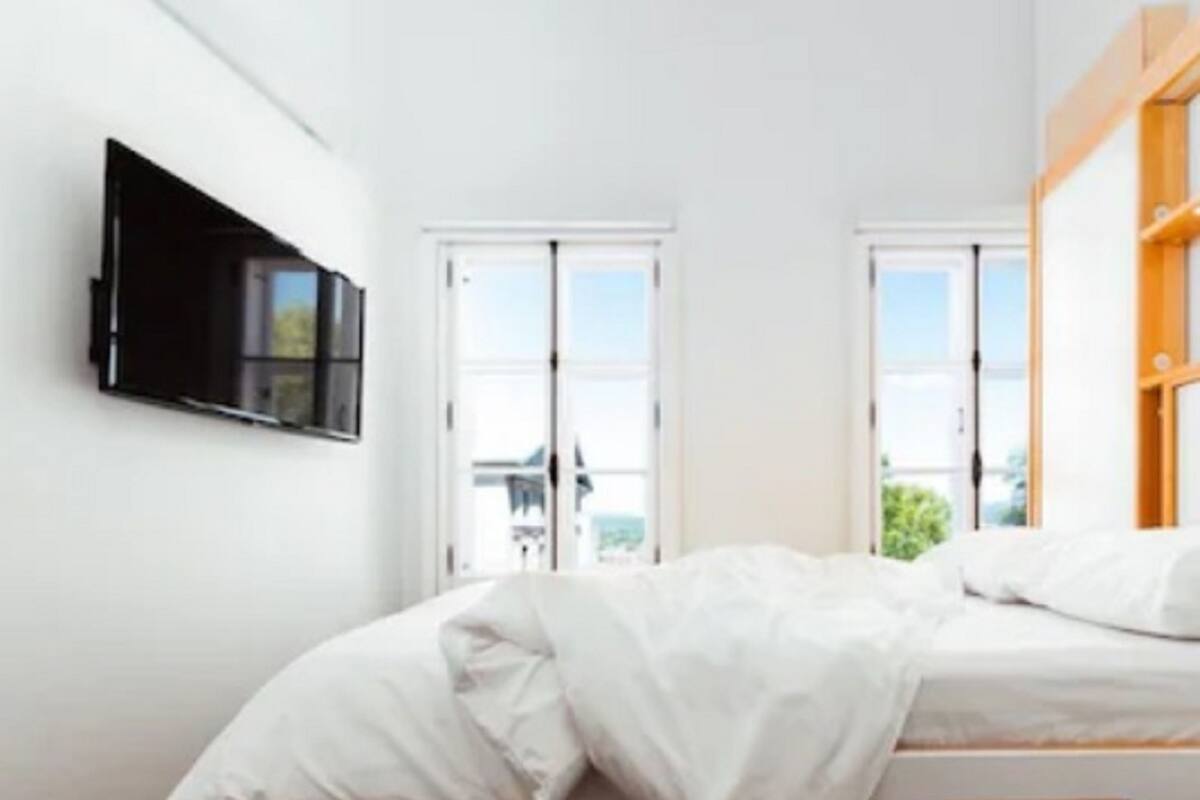 A bedroom with a bed, a TV mounted on the wall, and two windows.